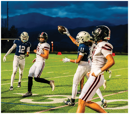 Four people in football uniforms are on a field at dusk, with one person in a blue jersey catching a football while others in white and blue jerseys watch or move nearby. Description generated by AI