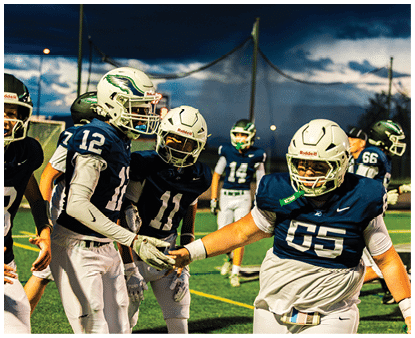Several people wearing football uniforms and helmets are on a field at dusk, with two of them engaging in a handshake or hand gesture. Description generated by AI
