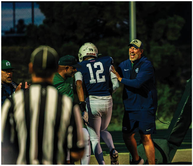 An adult in sportswear excitedly interacts with a person in a blue football uniform near the sideline while another person in a white football uniform walks nearby and a referee stands in the foreground. Description generated by AI