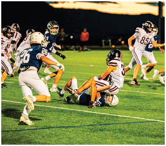 Players in football uniforms are actively engaged in a nighttime game on a well-lit field with one player tackling another on the ground. Description generated by AI