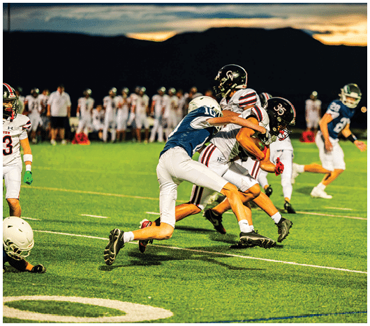 Two people in football uniforms are actively tackling another player holding a football on a field during a game at sunset. Description generated by AI