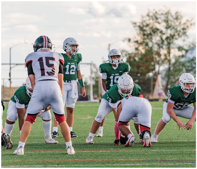 Two people wearing football uniforms are on a field with one person in a green jersey running towards the other person in a white jersey against a backdrop of mountains and a partly cloudy sky. Description generated by AI