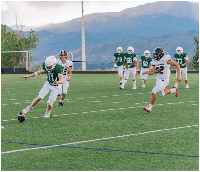 Two people wearing football uniforms are on a field with one person in a green jersey running towards the other person in a white jersey against a backdrop of mountains and a partly cloudy sky. Description generated by AI
