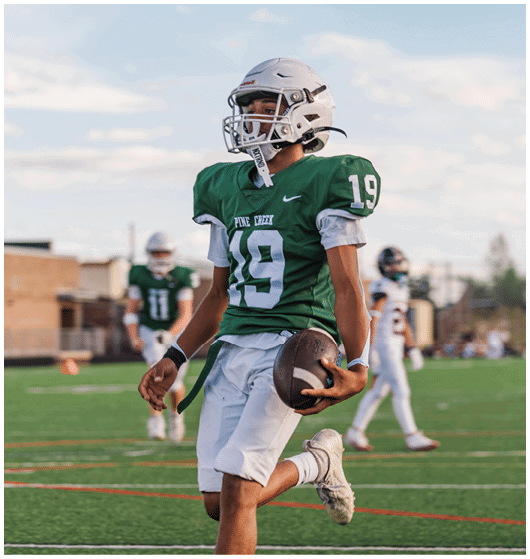 Two people wearing football uniforms are on a field with one person in a green jersey running towards the other person in a white jersey against a backdrop of mountains and a partly cloudy sky. Description generated by AI