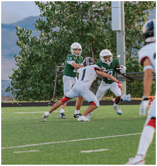 Two people wearing football uniforms are on a field with one person in a green jersey running towards the other person in a white jersey against a backdrop of mountains and a partly cloudy sky. Description generated by AI