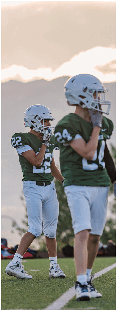 Two people wearing football uniforms are on a field with one person in a green jersey running towards the other person in a white jersey against a backdrop of mountains and a partly cloudy sky. Description generated by AI