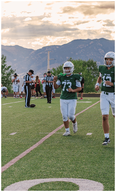 Two people wearing football uniforms are on a field with one person in a green jersey running towards the other person in a white jersey against a backdrop of mountains and a partly cloudy sky. Description generated by AI