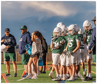 Two people wearing football uniforms are on a field with one person in a green jersey running towards the other person in a white jersey against a backdrop of mountains and a partly cloudy sky. Description generated by AI