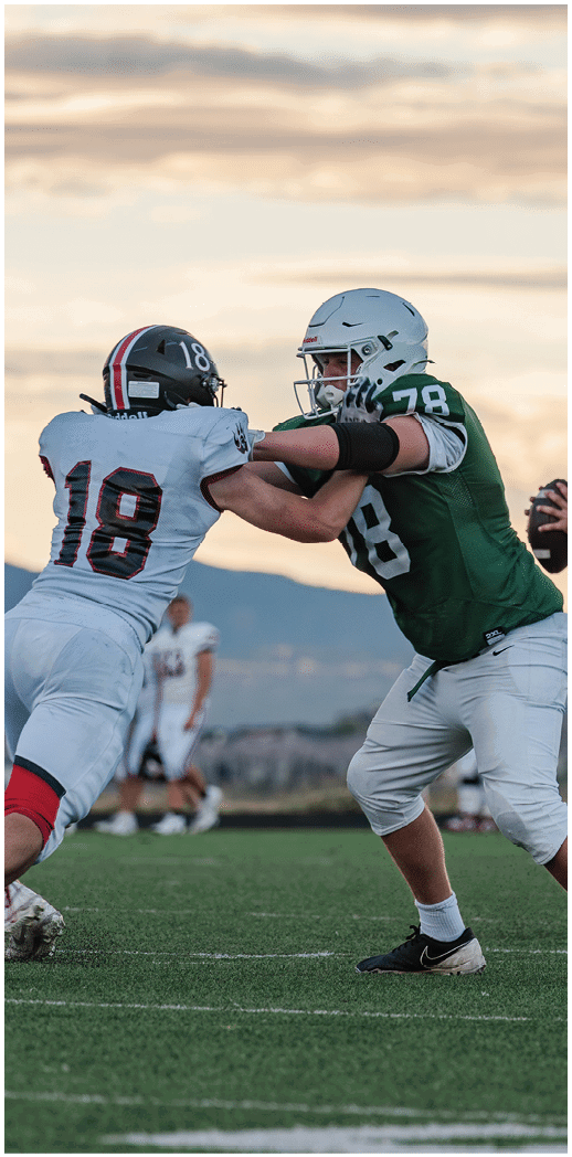 Two people wearing football uniforms are on a field with one person in a green jersey running towards the other person in a white jersey against a backdrop of mountains and a partly cloudy sky. Description generated by AI