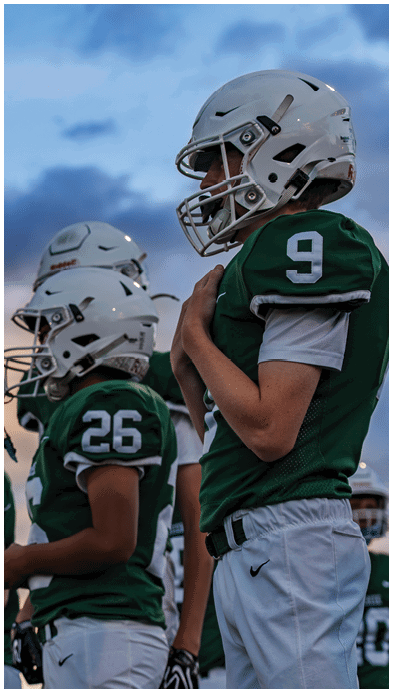 Two people wearing football uniforms are on a field with one person in a green jersey running towards the other person in a white jersey against a backdrop of mountains and a partly cloudy sky. Description generated by AI