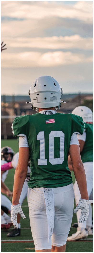 Two people wearing football uniforms are on a field with one person in a green jersey running towards the other person in a white jersey against a backdrop of mountains and a partly cloudy sky. Description generated by AI