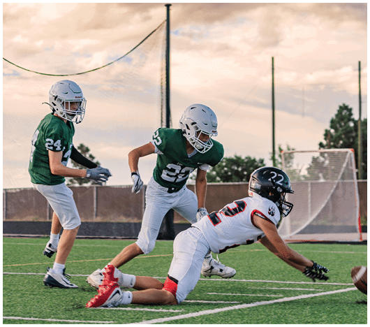 Five people in football uniforms stand on a field facing three other people in green and white football uniforms with helmets under a blue sky. Description generated by AI