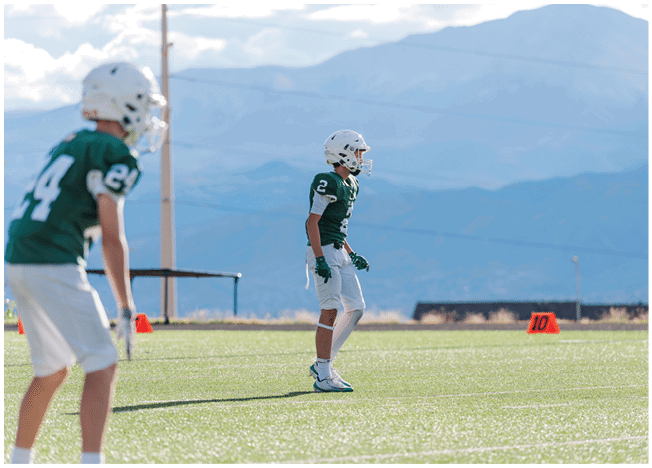 Five people in football uniforms stand on a field facing three other people in green and white football uniforms with helmets under a blue sky. Description generated by AI