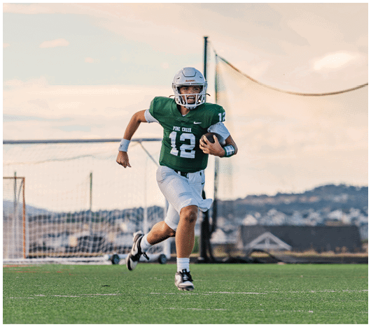 Five people in football uniforms stand on a field facing three other people in green and white football uniforms with helmets under a blue sky. Description generated by AI