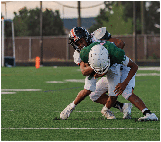 Five people in football uniforms stand on a field facing three other people in green and white football uniforms with helmets under a blue sky. Description generated by AI
