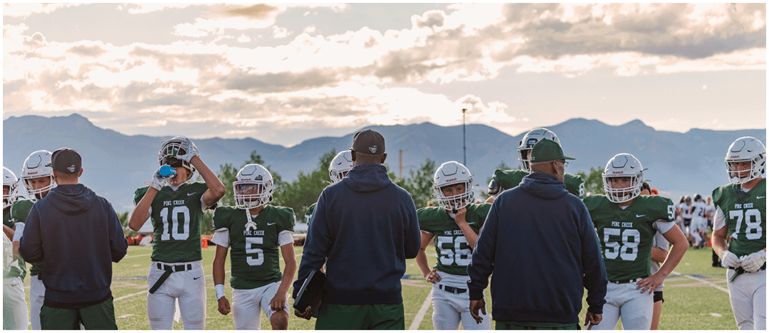 Five people in football uniforms stand on a field facing three other people in green and white football uniforms with helmets under a blue sky. Description generated by AI