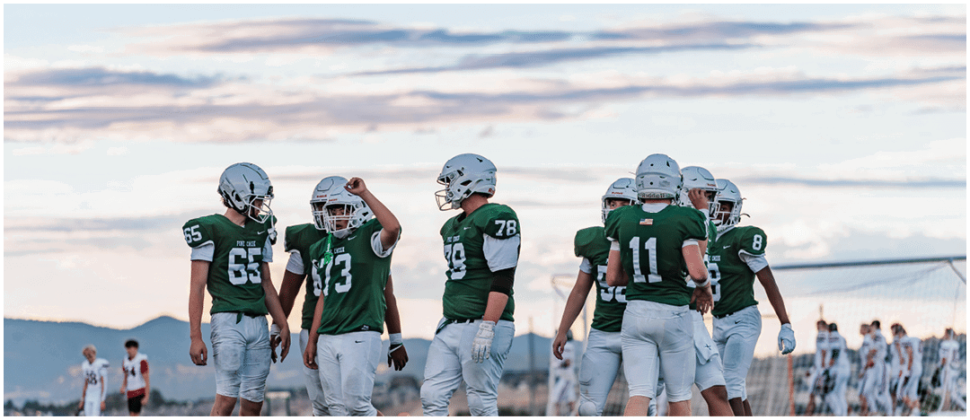 Five people in football uniforms stand on a field facing three other people in green and white football uniforms with helmets under a blue sky. Description generated by AI