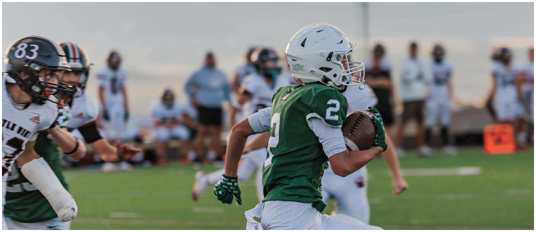 Five people in football uniforms stand on a field facing three other people in green and white football uniforms with helmets under a blue sky. Description generated by AI
