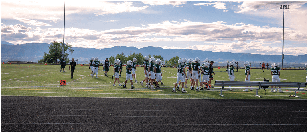 Five people in football uniforms stand on a field facing three other people in green and white football uniforms with helmets under a blue sky. Description generated by AI