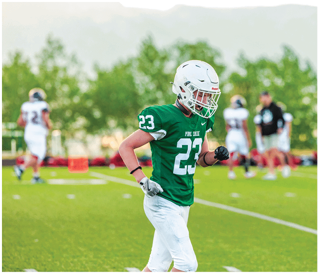Two people wearing football uniforms are on a field with one person in a green jersey running towards the other person in a white jersey against a backdrop of mountains and a partly cloudy sky. Description generated by AI