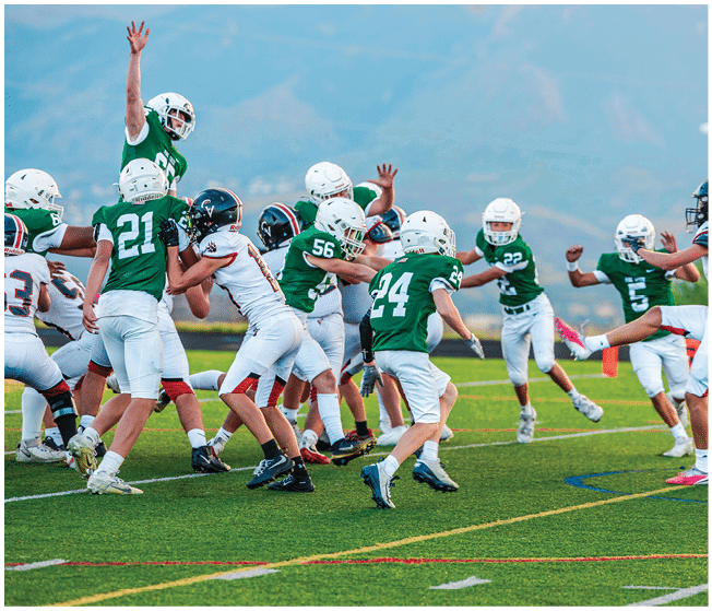 Two people wearing football uniforms are on a field with one person in a green jersey running towards the other person in a white jersey against a backdrop of mountains and a partly cloudy sky. Description generated by AI