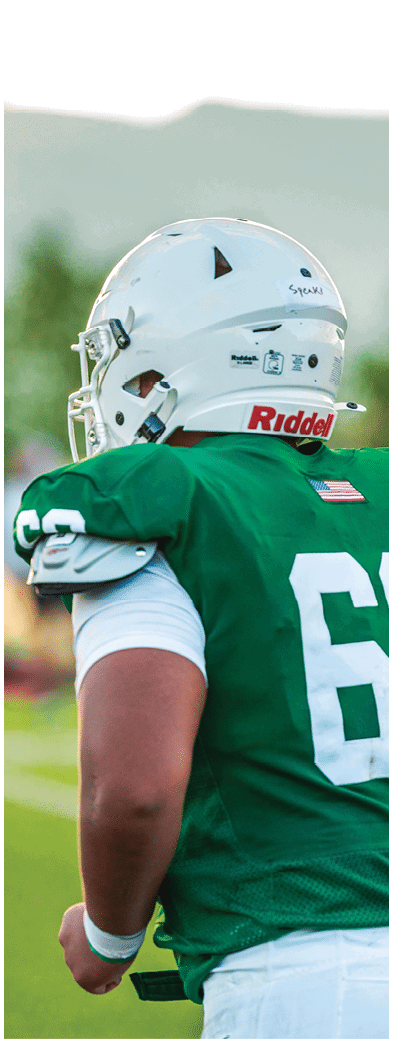 Two people wearing football uniforms are on a field with one person in a green jersey running towards the other person in a white jersey against a backdrop of mountains and a partly cloudy sky. Description generated by AI
