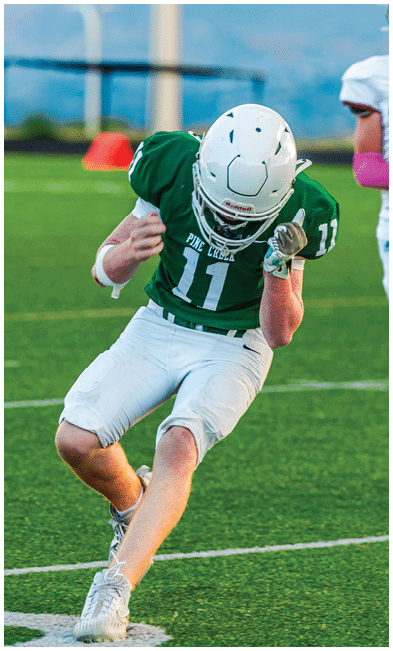 Two people wearing football uniforms are on a field with one person in a green jersey running towards the other person in a white jersey against a backdrop of mountains and a partly cloudy sky. Description generated by AI