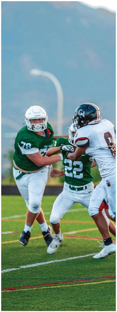 Two people wearing football uniforms are on a field with one person in a green jersey running towards the other person in a white jersey against a backdrop of mountains and a partly cloudy sky. Description generated by AI