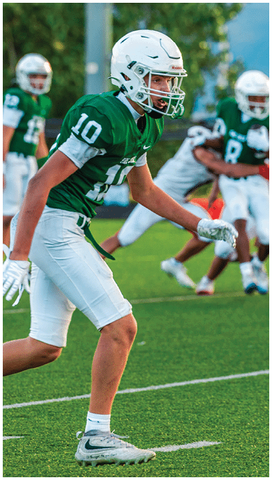 Two people wearing football uniforms are on a field with one person in a green jersey running towards the other person in a white jersey against a backdrop of mountains and a partly cloudy sky. Description generated by AI