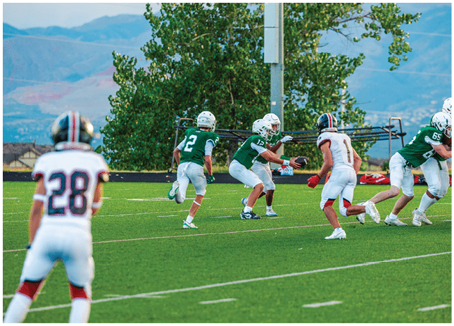 Five people in football uniforms stand on a field facing three other people in green and white football uniforms with helmets under a blue sky. Description generated by AI