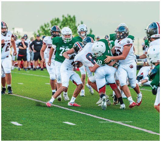 Five people in football uniforms stand on a field facing three other people in green and white football uniforms with helmets under a blue sky. Description generated by AI
