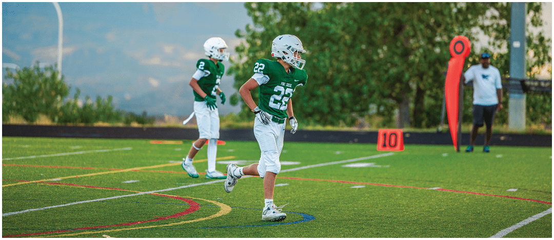 Five people in football uniforms stand on a field facing three other people in green and white football uniforms with helmets under a blue sky. Description generated by AI