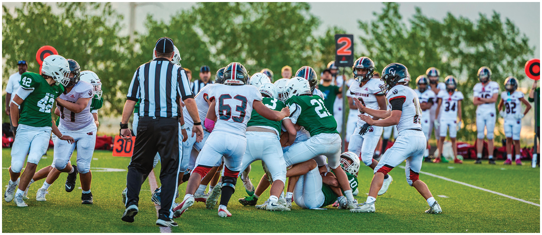 Five people in football uniforms stand on a field facing three other people in green and white football uniforms with helmets under a blue sky. Description generated by AI