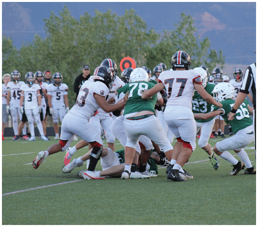 Several people in football uniforms are engaged in a play on a grassy field with others standing in the background near an orange yard marker. Description generated by AI