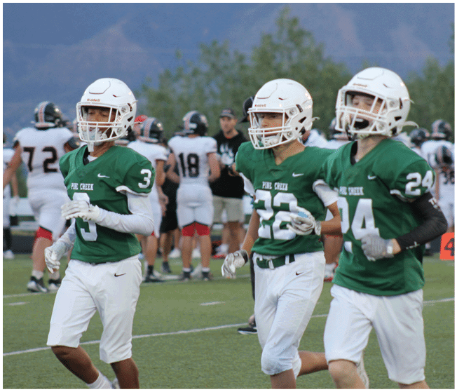 Three people wearing green football jerseys and white helmets are walking on a football field with other players and coaches in the background. Description generated by AI