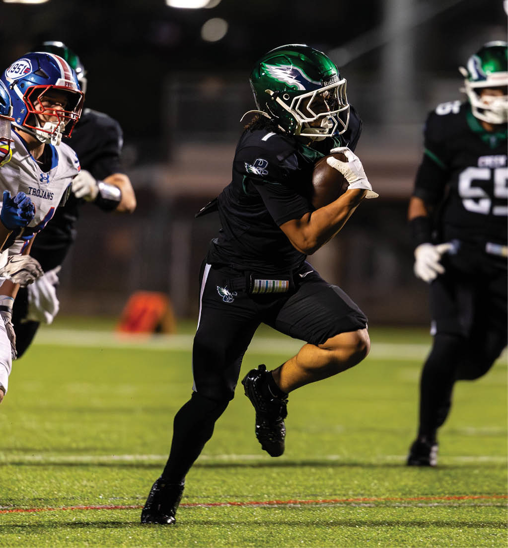 A person in a black football uniform with a green helmet is running with the ball while two people in white football uniforms with blue helmets chase behind on a grassy field. Description generated by AI
