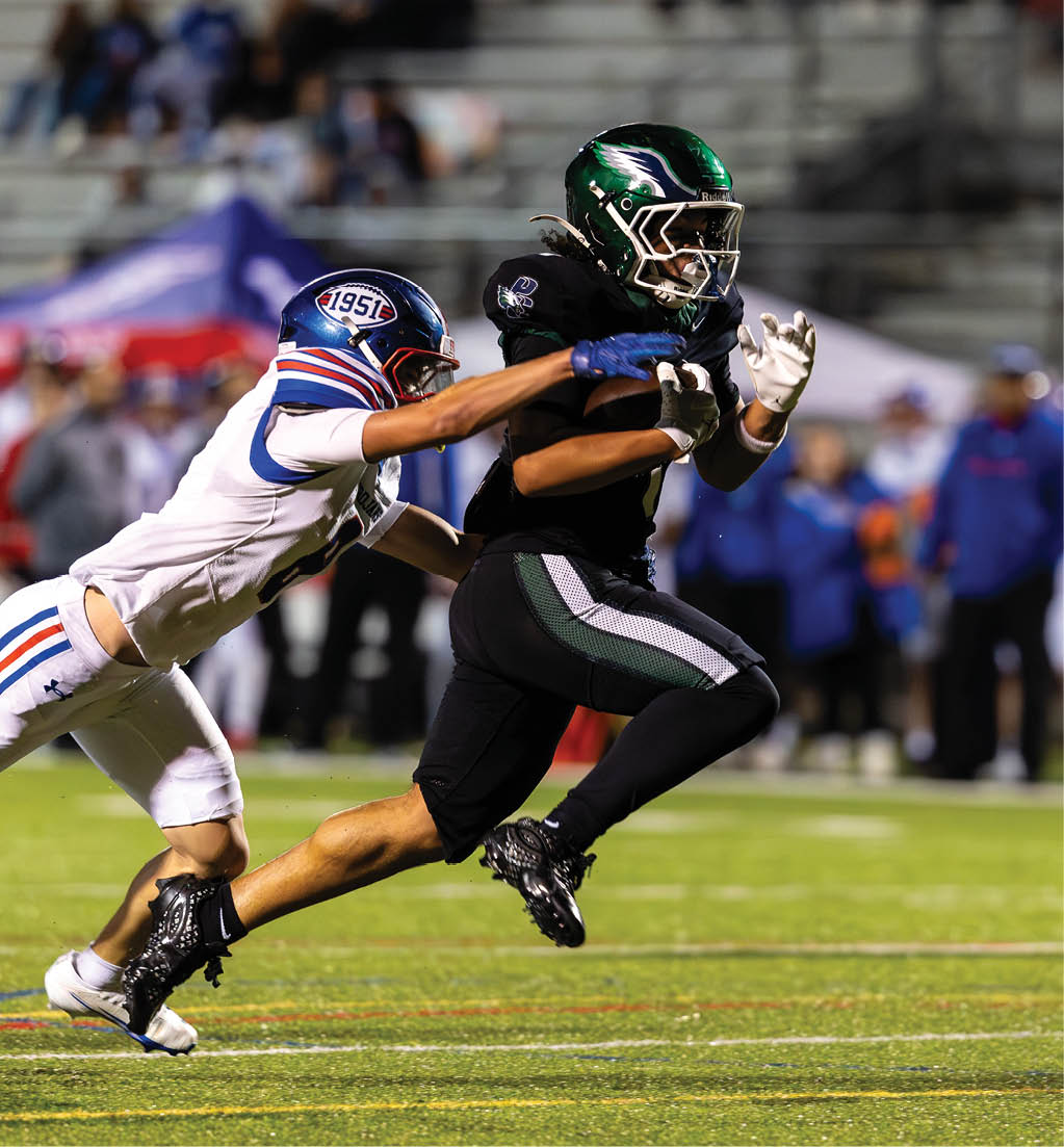 A person in black sports gear and a green helmet is running with a football while another person in white sports gear with blue and red stripes is attempting to tackle them on a football field. Description generated by AI
