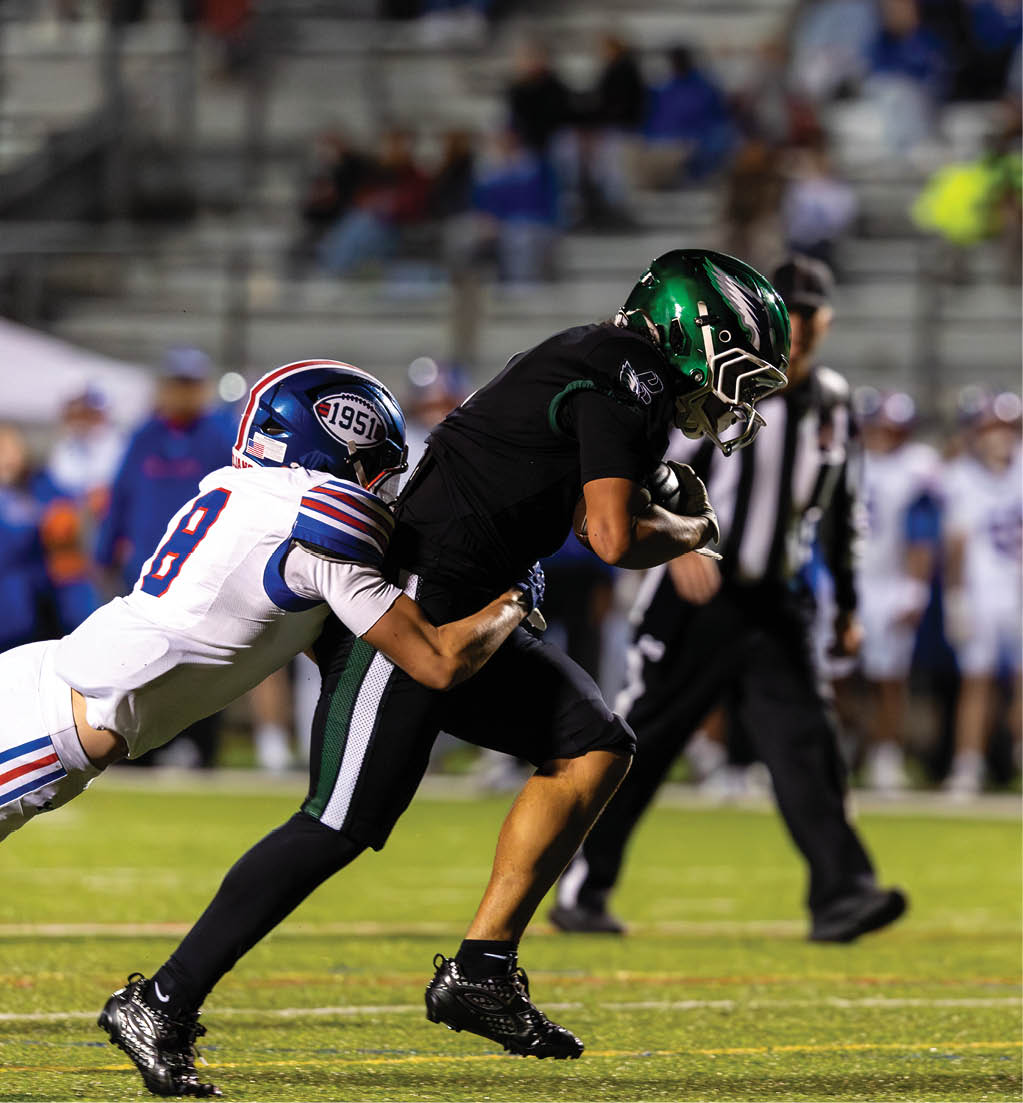 A person in a black football uniform with a green helmet is running while holding a football as another person in a white football uniform with blue and red stripes attempts to tackle them on a football field. Description generated by AI