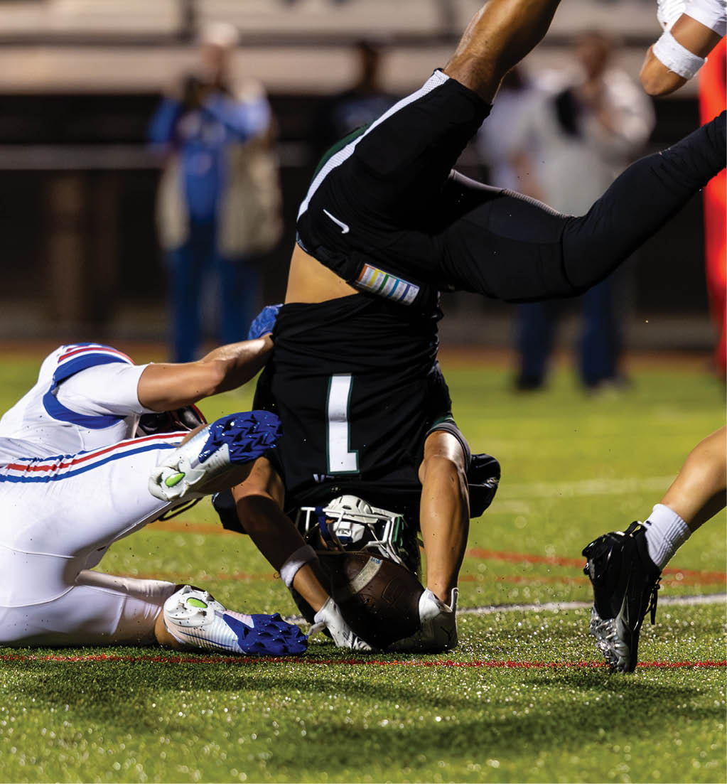 A person in a black sports uniform is upside down on a football field while two people in white uniforms are nearby, one on the ground and the other running. Description generated by AI