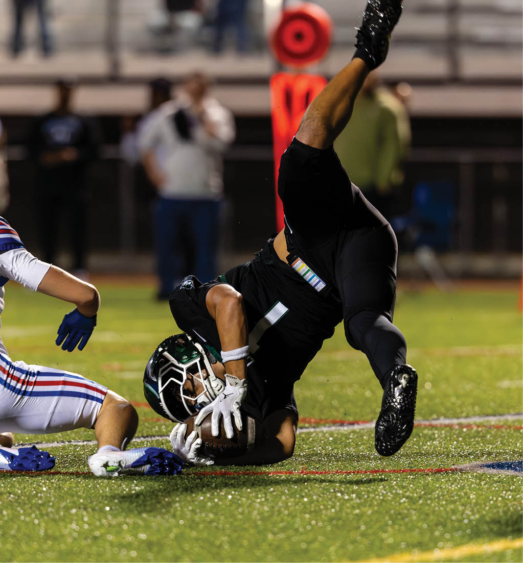 Two people in football uniforms are on the ground during a game, with one person holding the ball and the other sitting nearby on a grassy field. Description generated by AI