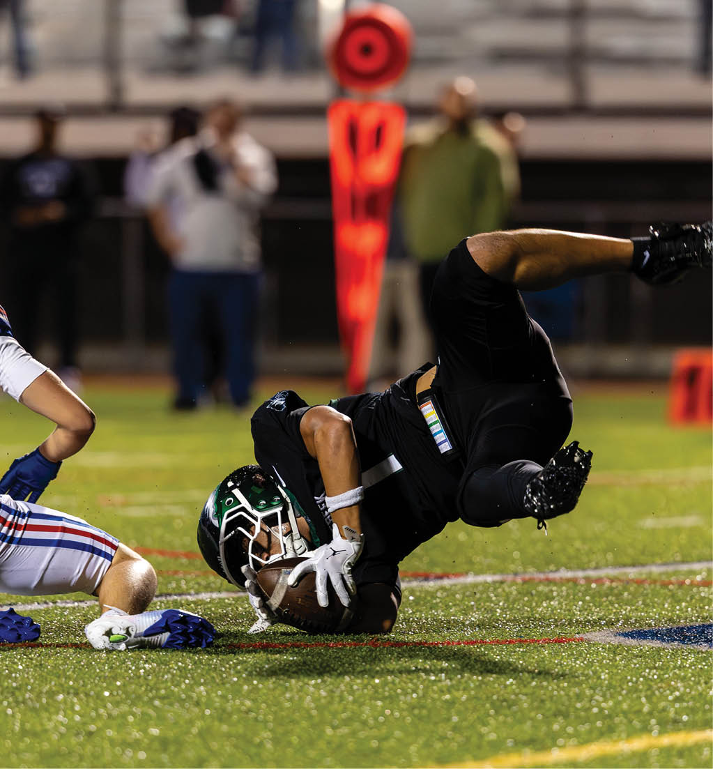 A person in black sports gear is falling forward while holding a football on a field as another person in white sports gear with the number 8 kneels nearby. Description generated by AI
