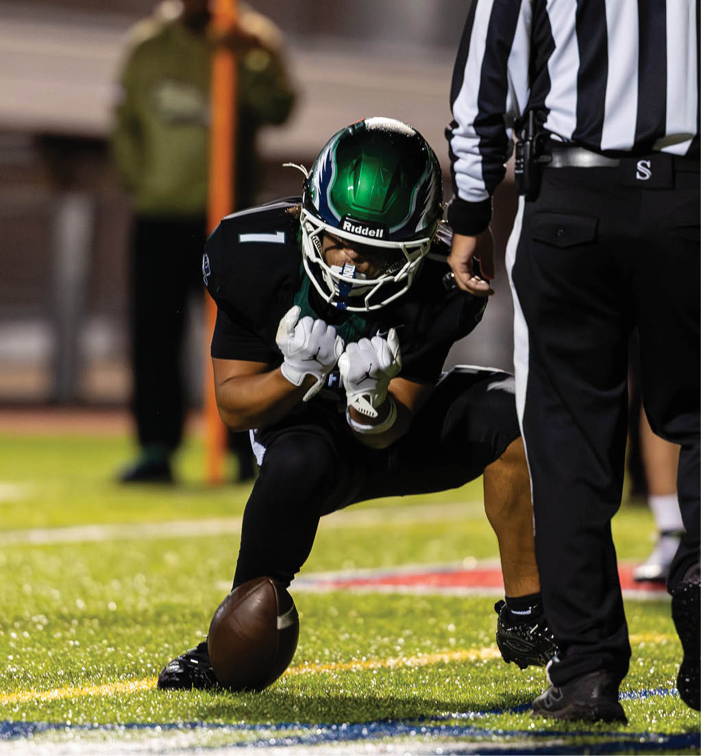 A person in a green helmet and black uniform is crouching on a football field, holding a football on the ground, while a referee stands nearby. Description generated by AI