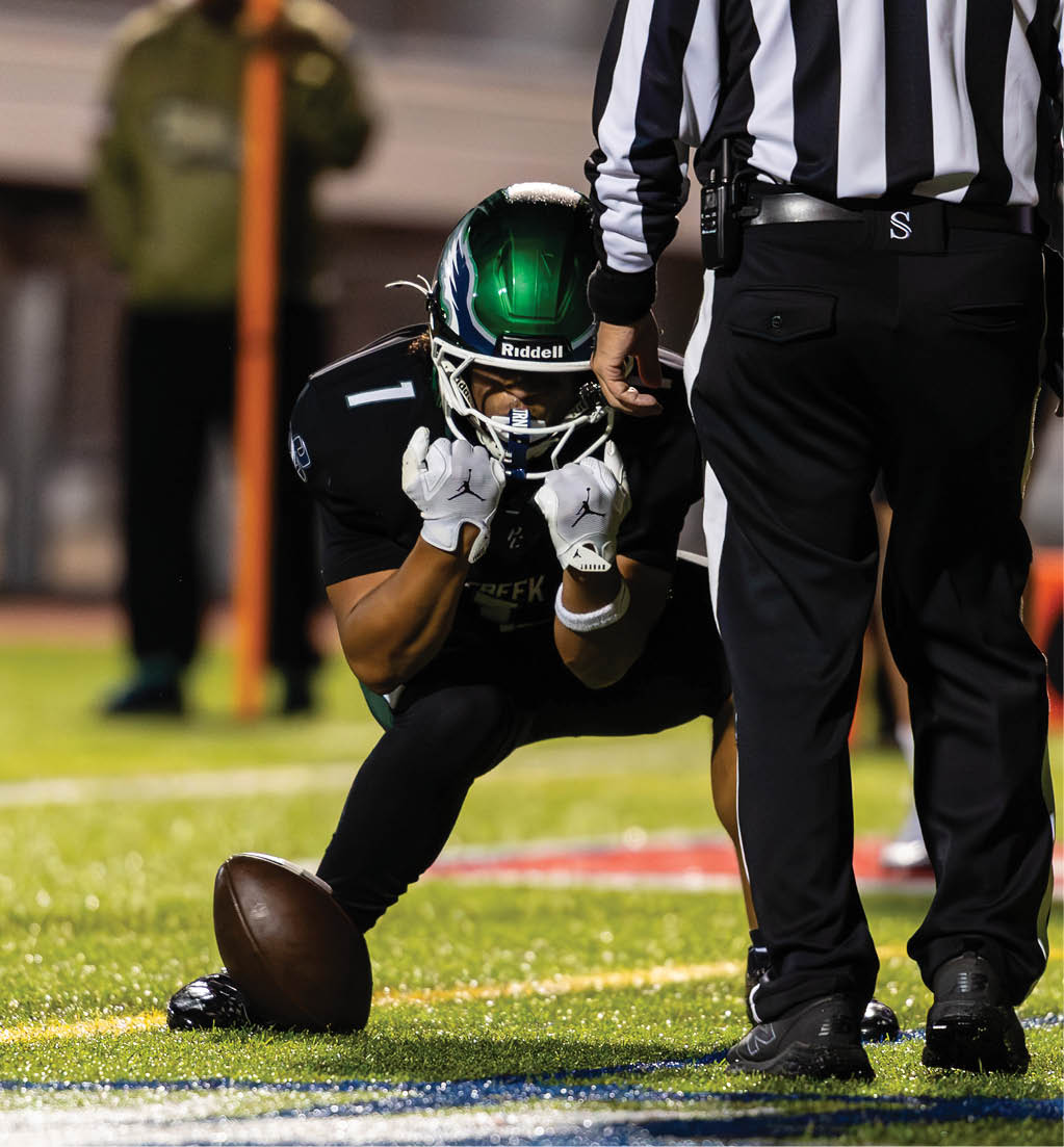 A person in a green helmet and black uniform is crouched on a football field holding a football upright while a referee in a black and white striped shirt stands nearby. Description generated by AI