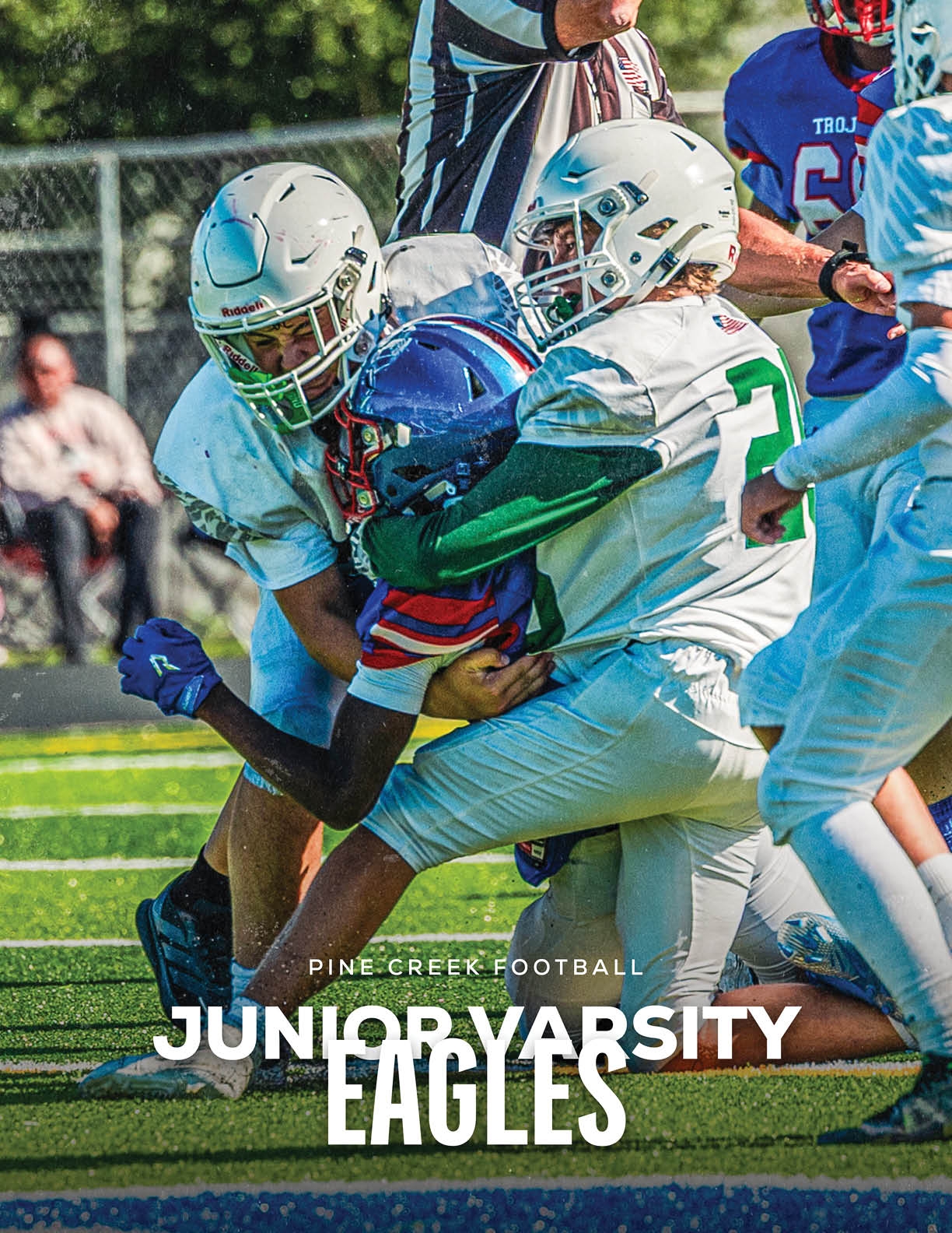 Two football players in white uniforms tackle a player in a blue and red uniform on a green field during a game. Description generated by AI