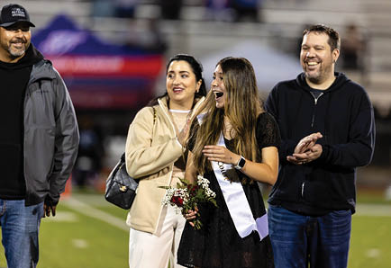 A person wearing a sash and holding flowers stands smiling and laughing on a sports field while three other people nearby clap and smile. Description generated by AI