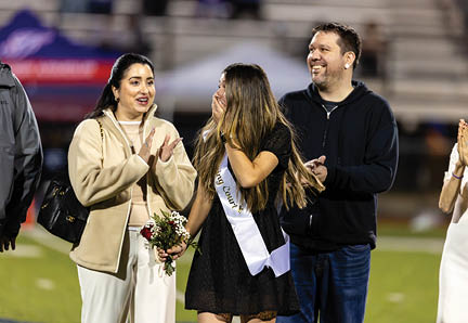 Three people stand on a field with one person wearing a sash and holding flowers while the others smile and clap. Description generated by AI