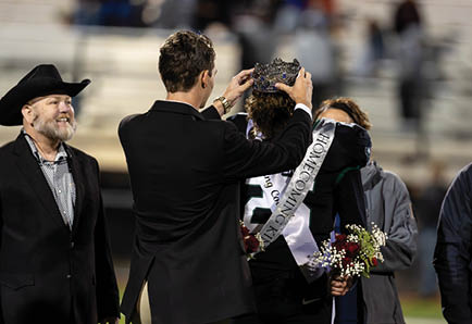 An adult in a suit places a crown on a person wearing a sash that reads “Homecoming King" while others stand nearby holding flowers in a stadium setting. Description generated by AI