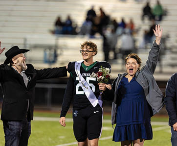 A person wearing a sports uniform and sash holds flowers while standing between two people, one raising a hand excitedly and the other reaching out, all on a sports field with blurred spectators in the background. Description generated by AI