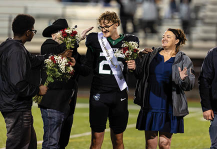 A person wearing a football uniform and a sash that says “Homecoming Court" is smiling and holding flowers while surrounded by three other people on a sports field. Description generated by AI