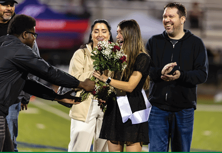 People on a sports field are smiling and presenting a bouquet of flowers to a person wearing a black dress and a white sash. Description generated by AI